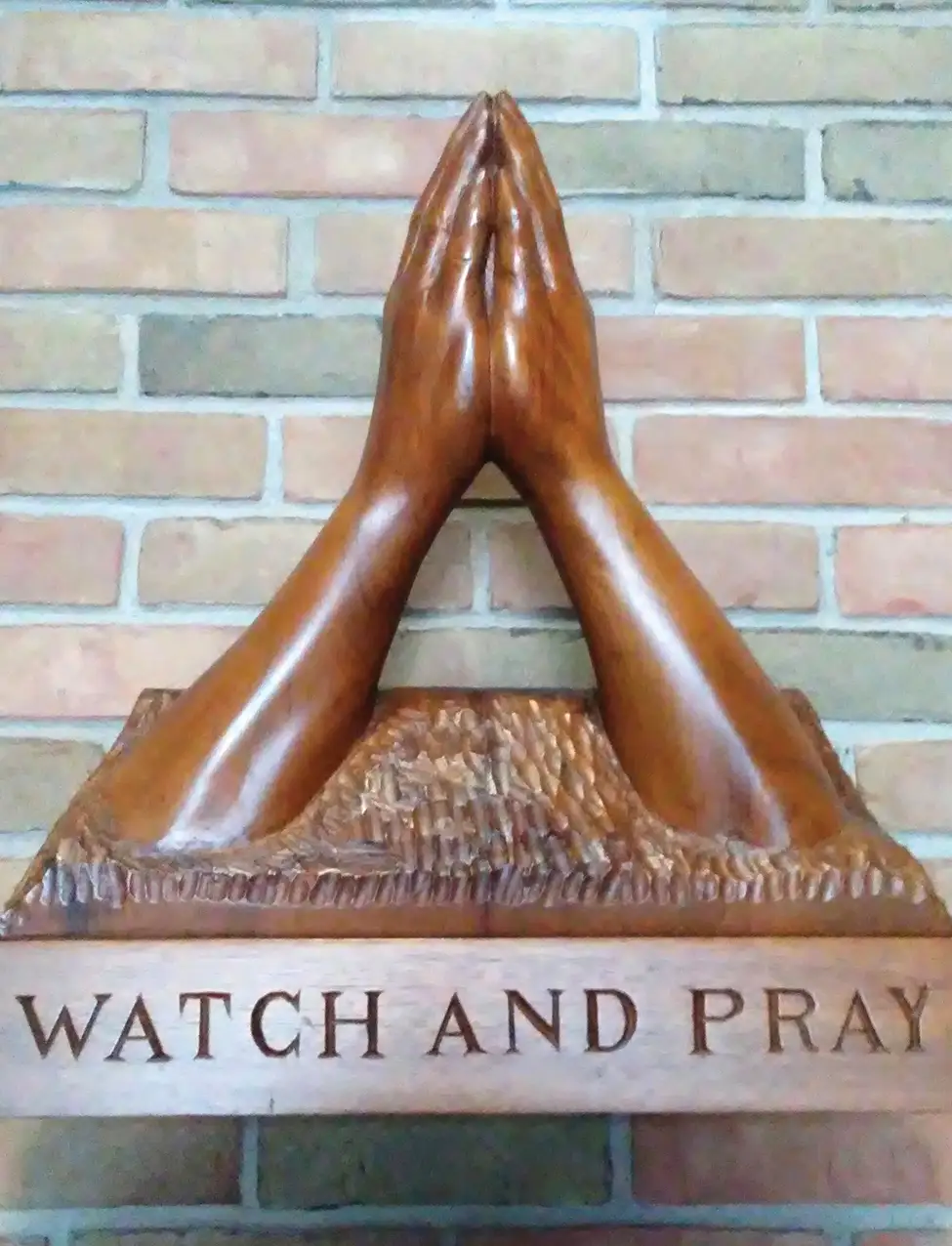 Wooden sculpture of praying hands on brick wall with the words watch and pray