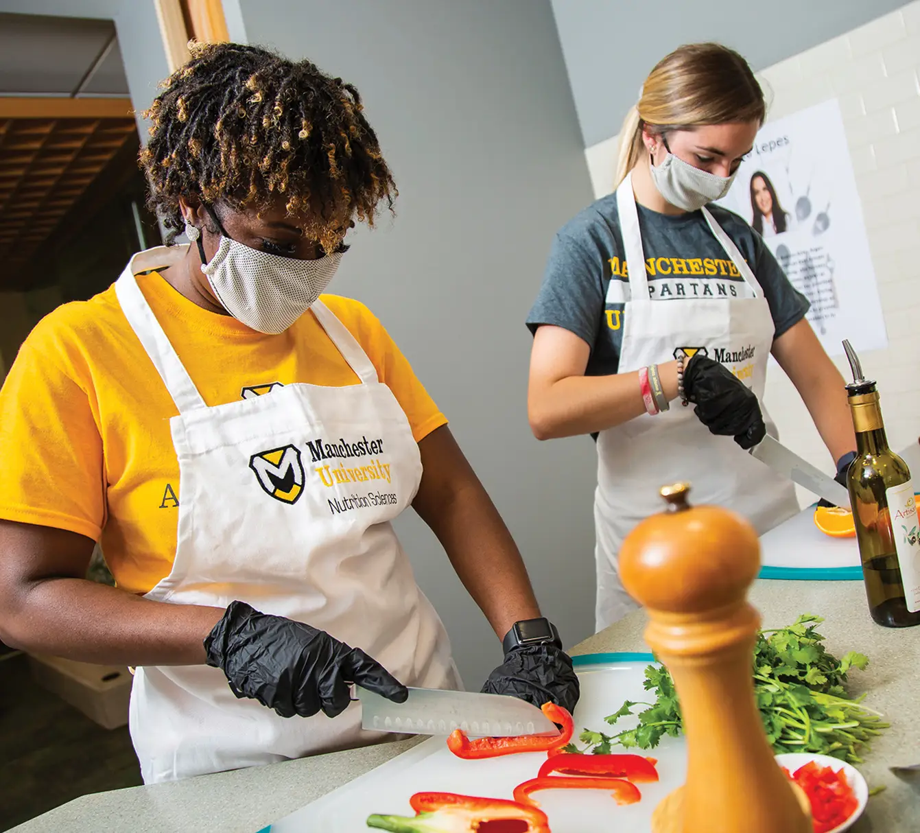 Two female students wearing black gloves and aprons cutting vegetables in a culinary class
