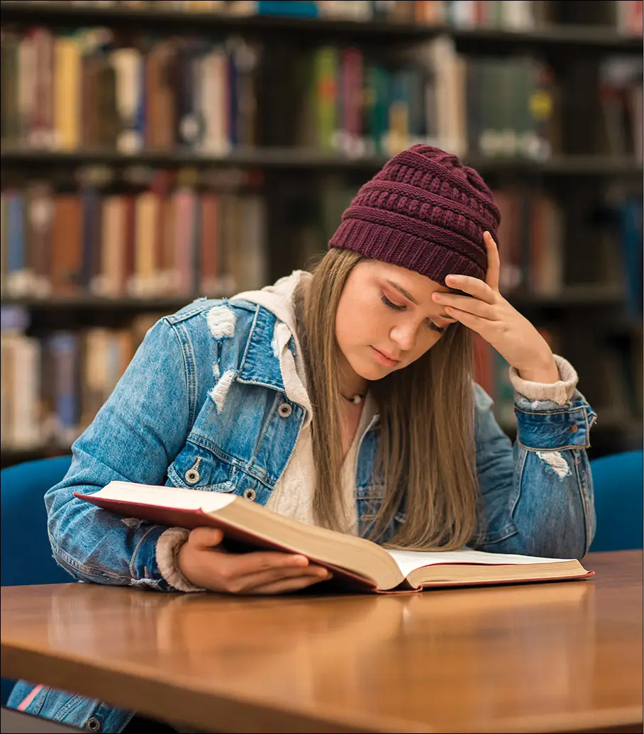 Girl with burgundy beanie and jean jacket sitting at table with hand on head reading book in library