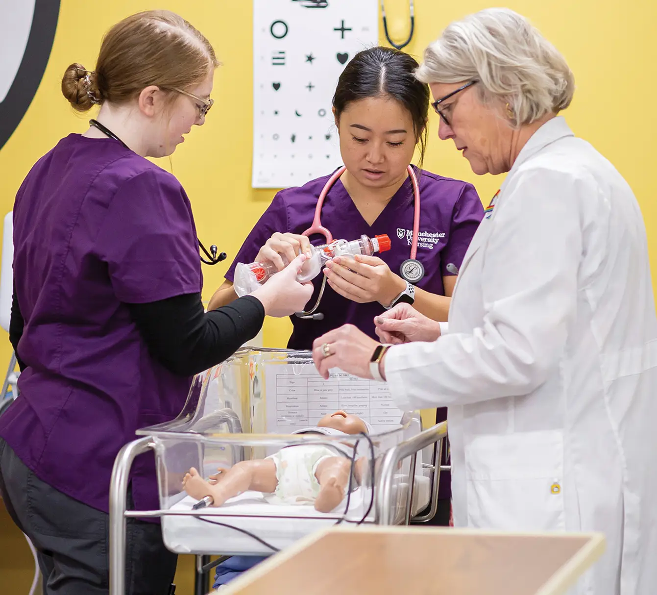 Two nursing students and an instructor standing over hospital bassinet with toy baby in it