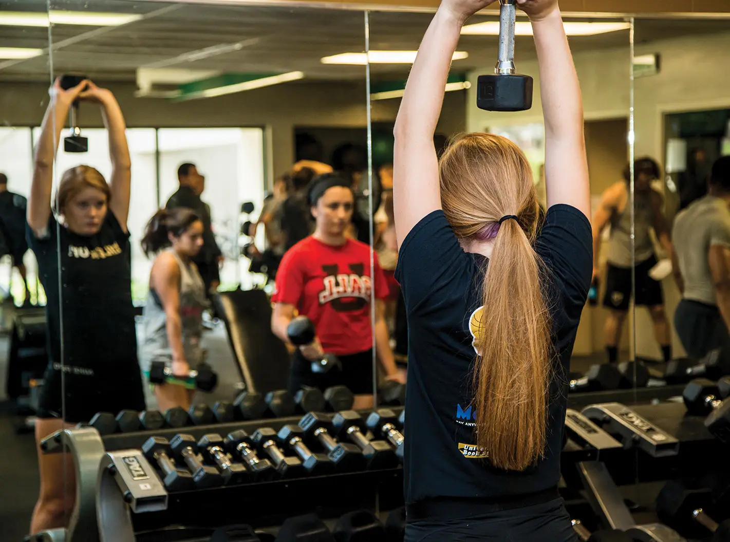 Female student with her back turned looking in mirror of weight room lifting dumb bell over her head