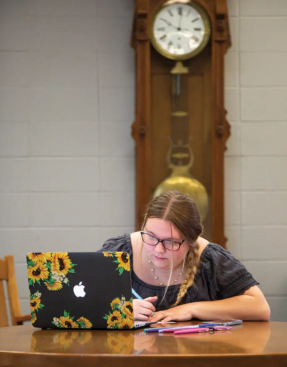 Student writing on paper while looking at laptop with sunflowers on it and a large grandfather clock in the background