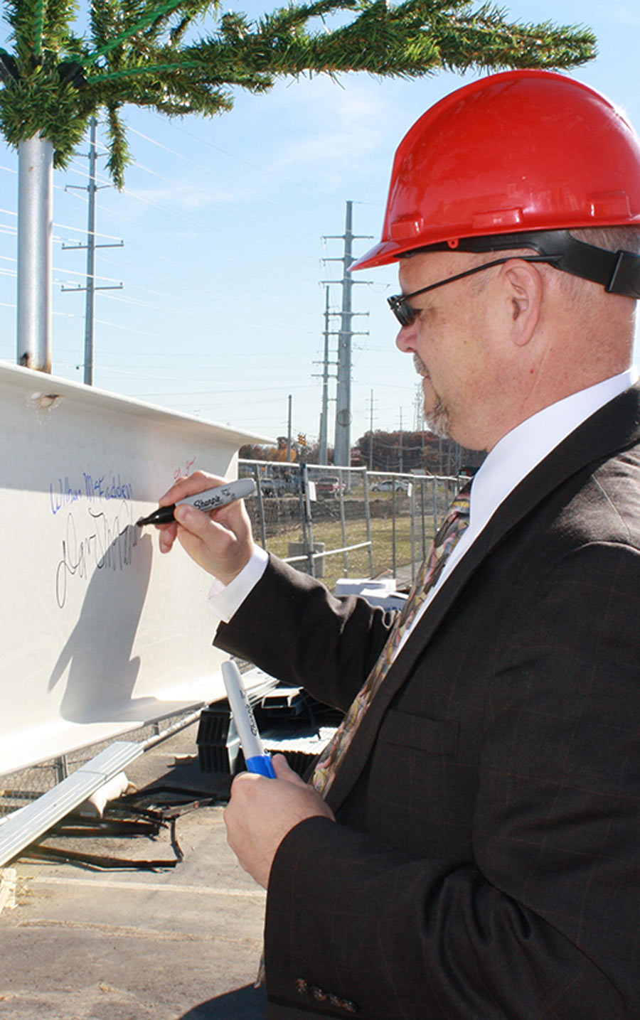Dave McFadden in a black suit and red hard hat, signing a metal beam that is being used for construction