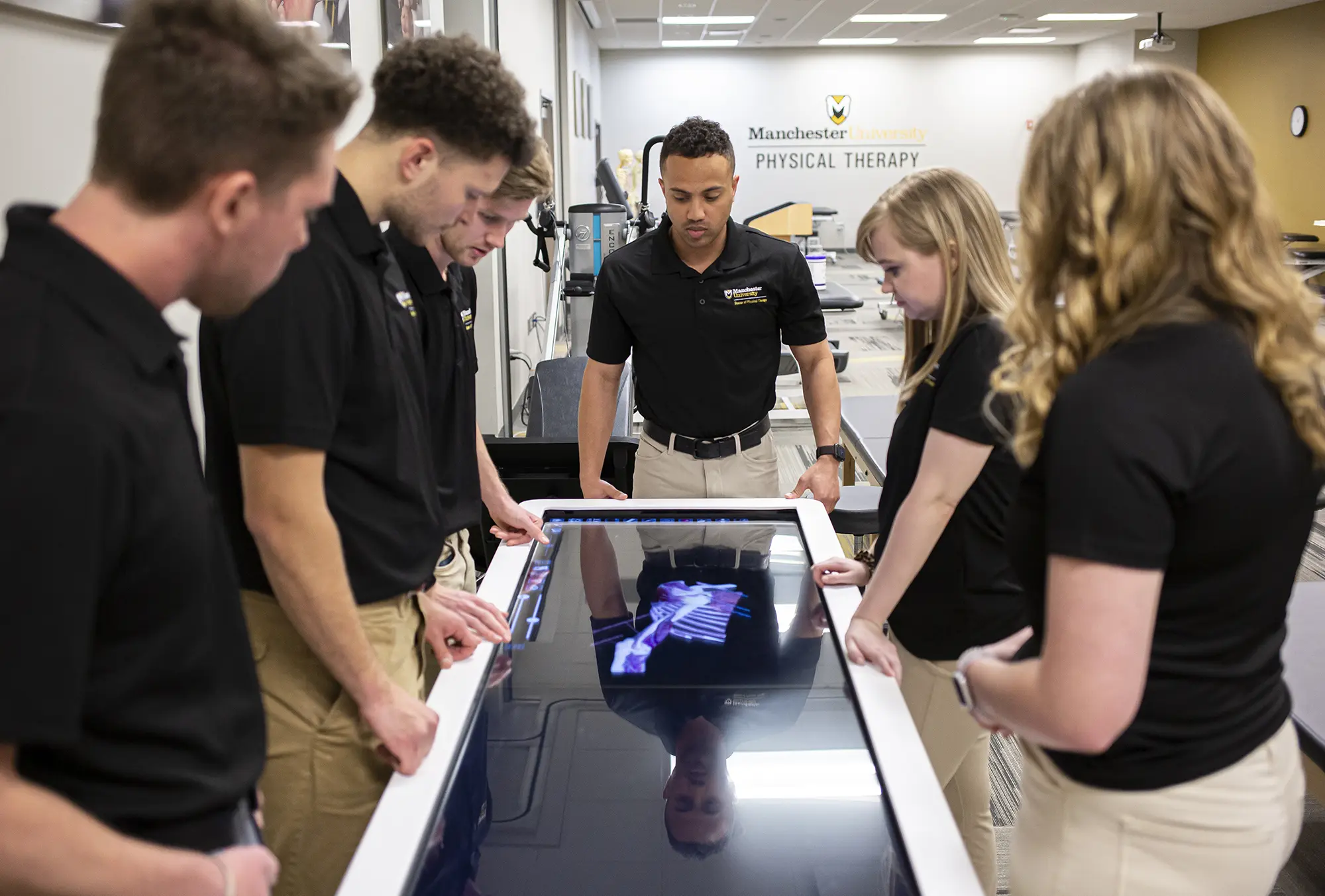 Six Manchester University Physical Therapy students (four men plus two women) inside a room glance downward at a black touchscreen that displays a three-dimensional x-ray scan of a body part with other helpful beneficial informational icon buttons on the side and at the bottom of the black touchscreen
