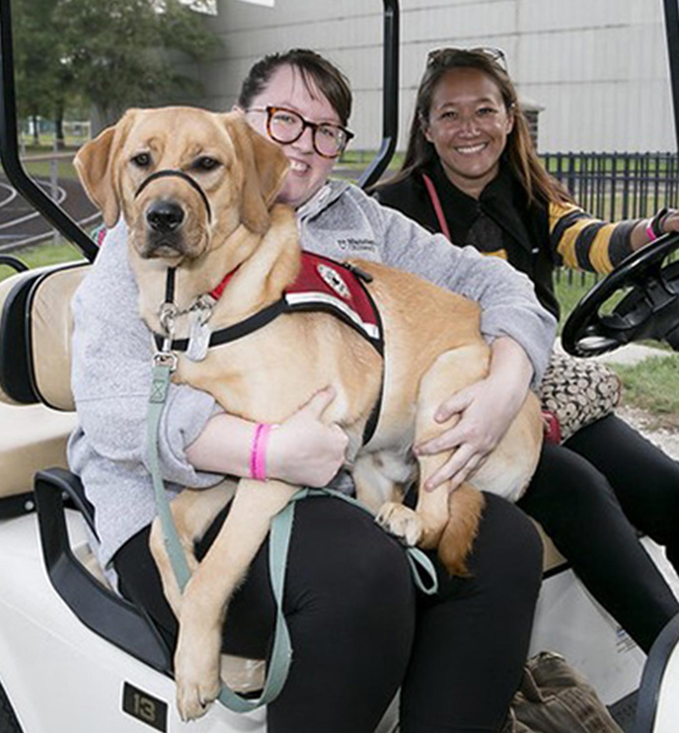 two women sit in a golf cart smiling, while one holds the large golden Labrador Neelix in their lap